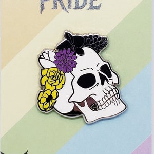Puede incluir: Un pin de esmalte blanco y negro con una calavera que lleva una corona de flores. Las flores son moradas, amarillas y blancas. El pin dice "Spooky Pride" y "ArtByKino.com" en la tarjeta.