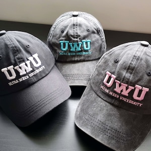 以下が含まれることがあります： 刺繍されたテキストが入った3つのグレーの野球帽。「UWU」と「ULTRA WEEB UNIVERSITY」の文字が白、水色、ピンクで表示されています。帽子は柔らかく、使い古された生地で作られています。