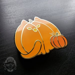 May include: Gold enamel pin featuring two orange cats shaped like pumpkins, with one cat holding a smaller orange pumpkin. The pin has a glittery finish.