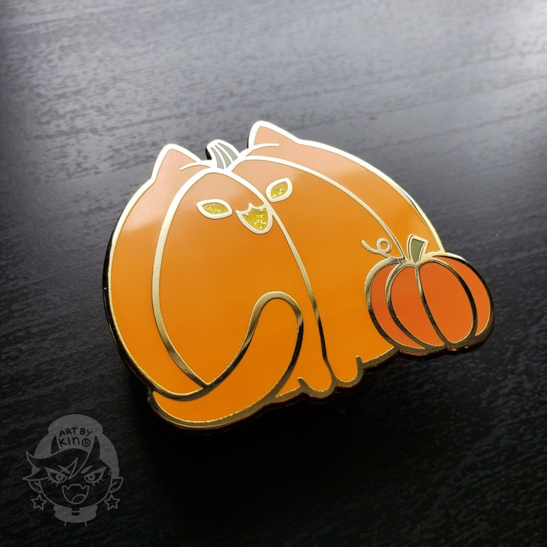 May include: Gold enamel pin featuring two orange cats shaped like pumpkins, with one cat holding a smaller orange pumpkin. The pin has a glittery finish.