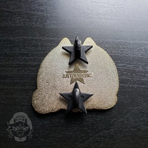 May include: Gold-colored metal pin with a black cat silhouette. The back of the pin has three black star-shaped fasteners and the text "ART BY KINO".