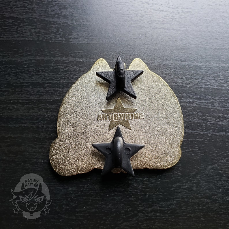 May include: Gold-colored metal pin with a black cat silhouette. The back of the pin has three black star-shaped fasteners and the text "ART BY KINO".