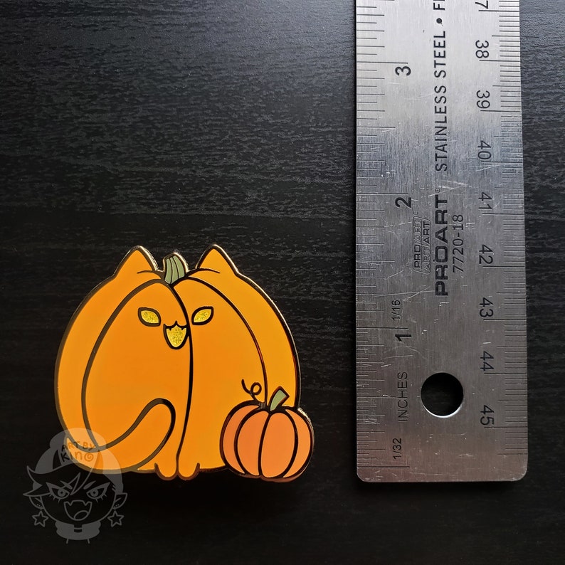 May include: Gold-colored enamel pin featuring a cat dressed as a pumpkin. The cat is wearing a pumpkin costume with a small pumpkin in its paw. The pin is approximately 1.5 inches tall.