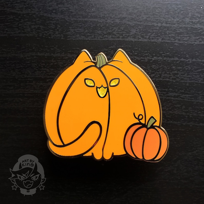 May include: A gold-colored enamel pin of a cat dressed as a pumpkin. The cat has orange fur, black eyes, and a black nose. The pumpkin has a green stem and a small pumpkin on the side.