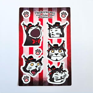 May include: A sheet of vinyl stickers featuring five different black cat faces with red accents and top hats. The stickers are on a red and white striped background with paw prints. The text "Huskermemes vinyl sticker sheet artbykino.com" is at the top of the sheet.