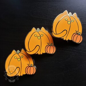 May include: Three gold enamel pins shaped like pumpkins with cat faces. Each pin has a smaller pumpkin on the side. The pumpkins are orange with black eyes and a black mouth.