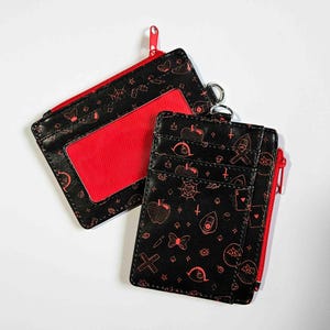 May include: Two black cardholders with red zips and accents. One has a clear window, the other has card slots. Both are decorated with red illustrations of apples, spiders, and other gothic-themed designs.