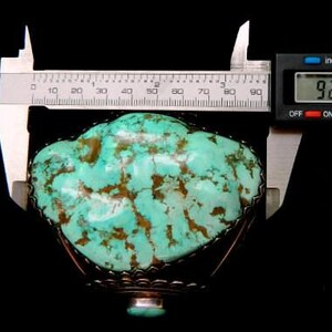 以下が含まれることがあります： 茶色のマトリックスが入ったターコイズの石が、装飾的な金属フレームにセットされています。石はデジタルノギスで測定され、幅は234.6 cmです。背景は黒です。