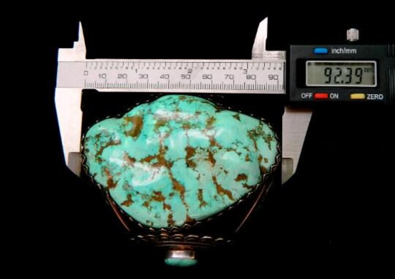 以下が含まれることがあります： 茶色のマトリックスが入ったターコイズの石が、装飾的な金属フレームにセットされています。石はデジタルノギスで測定され、幅は234.6 cmです。背景は黒です。