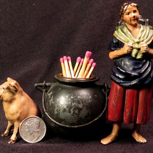May include: A vintage match holder featuring a woman figurine, a pig figurine, and a black cauldron with a lid. The cauldron is filled with matches with pink tips.