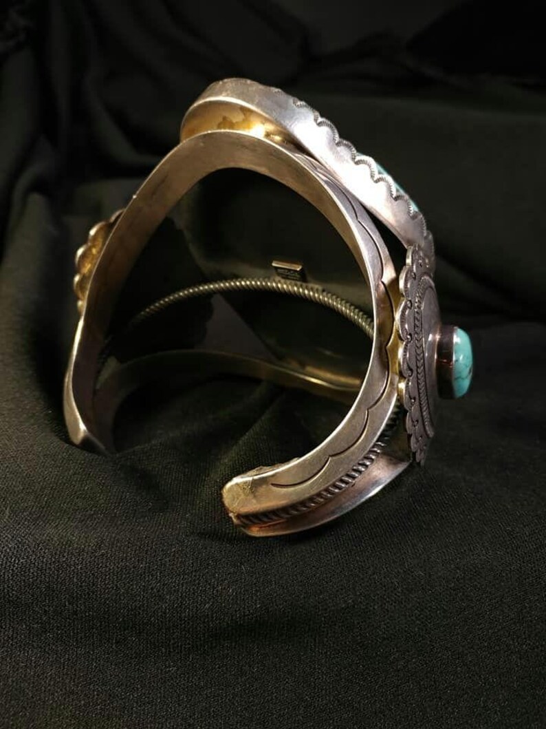 以下が含まれることがあります： ターコイズのアクセントが付いたシルバーのカフブレスレット。ブレスレットは、装飾的なエッジや中央のターコイズストーンなど、複雑な金属細工が特徴です。デザインは、南西部またはネイティブアメリカンの美学を持っています。