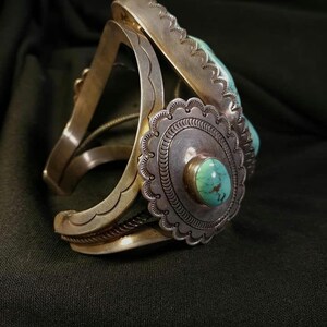 以下が含まれることがあります： 中央にターコイズストーンをあしらったシルバーのカフブレスレット。ブレスレットは石の周りに装飾的な花の模様があり、端に刻印された模様があります。カフはオープンで、ヴィンテージまたは南西部のスタイルです。