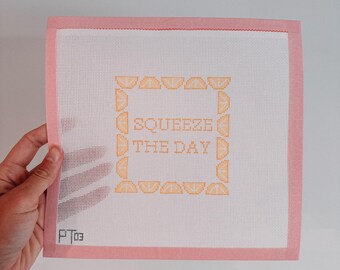 May include: A white canvas with a pink border, featuring a cross-stitch design of orange slices framing the text "SQUEEZE THE DAY". The canvas is signed "P Too".