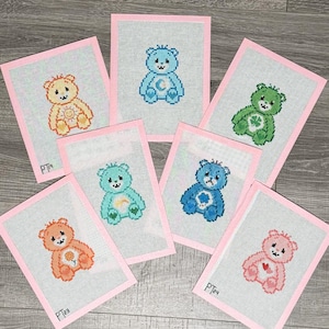 May include: Six framed cross-stitch embroidery designs featuring Care Bear characters. Each design is on a light blue fabric with a pink border. The characters include a bear with a sun, a bear with a moon, a bear with a rainbow, a bear with a clover, a bear with a heart, and a bear with a flower.