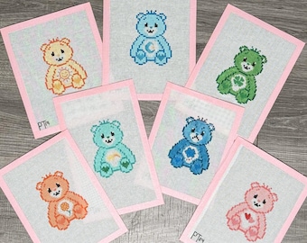 May include: Six framed cross-stitch embroidery designs featuring Care Bear characters. Each design is on a light blue fabric with a pink border. The characters include a bear with a sun, a bear with a moon, a bear with a rainbow, a bear with a clover, a bear with a heart, and a bear with a flower.