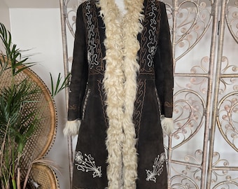 May include: A dark brown suede coat with cream-colored faux fur trim and lining. The coat features embroidered floral and swirl designs in white and brown. The coat is displayed on a wooden mannequin.