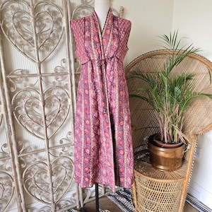 Puede incluir: Vestido largo sin mangas sobre un maniquí. El vestido presenta un estampado floral rosa con rayas verticales y un lazo en la cintura. El vestido se exhibe frente a una pantalla decorativa y una silla de mimbre.