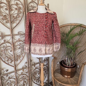Puede incluir: Chaqueta vintage roja con estampado floral, mangas largas y cierre de botones. La chaqueta presenta un estampado floral crema en el cuello y el dobladillo. La chaqueta se exhibe en un maniquí de madera.