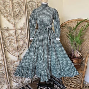 May include: A long-sleeved, floor-length dress in a dark green and white houndstooth pattern. The dress features a high neck with white lace trim, puffed sleeves with lace-trimmed cuffs, a cinched waist with a matching tie belt, and a ruffled hem.