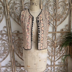 Puede incluir: Un chaleco sin mangas de color crema con ribete negro y bordado floral intrincado en tonos marrones, naranjas y negros. El chaleco tiene cierre de botones y se muestra en un maniquí.