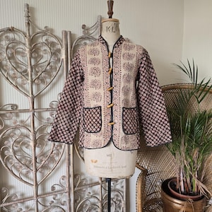 Puede incluir: Chaqueta estampada en beige y negro, con cuello mandarín y botones de madera. La chaqueta, expuesta en un maniquí, tiene dos bolsillos delanteros. Las mangas y la espalda están estampadas con un pequeño diseño floral.