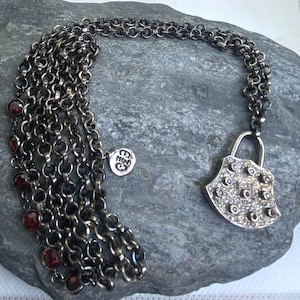May include: A silver chain necklace with a padlock pendant. The padlock is decorated with small red gemstones and clear rhinestones. The chain is layered and has small red gemstones hanging from it. The necklace has a small silver tag with the word "Nina" on it.