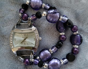 May include: A silver watch with a white face and black hands. The watch is attached to a purple and black beaded bracelet. The watch face has the words "Geneve Quartz" printed on it.