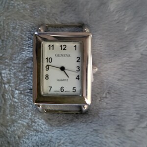 May include: A silver rectangular wristwatch with a white face. The watch face has black numbers and hands, and the text "GENEVA QUARTZ" is printed on the face.