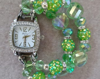 May include: A silver-toned wristwatch with a square face, surrounded by small, clear crystals. The watch face is white with black numerals and hands. The watch is paired with a double strand bracelet of green beads in various shapes and sizes.