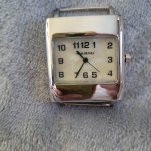 May include: A silver watch with a white face and black numbers. The watch has a square face and a silver band.