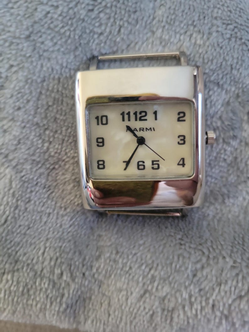 May include: A silver watch with a white face and black numbers. The watch has a square face and a silver band.
