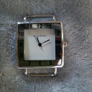 May include: A silver square watch with a white face and black numbers. The watch has a green border around the face and the word "WARMI" printed on the face.