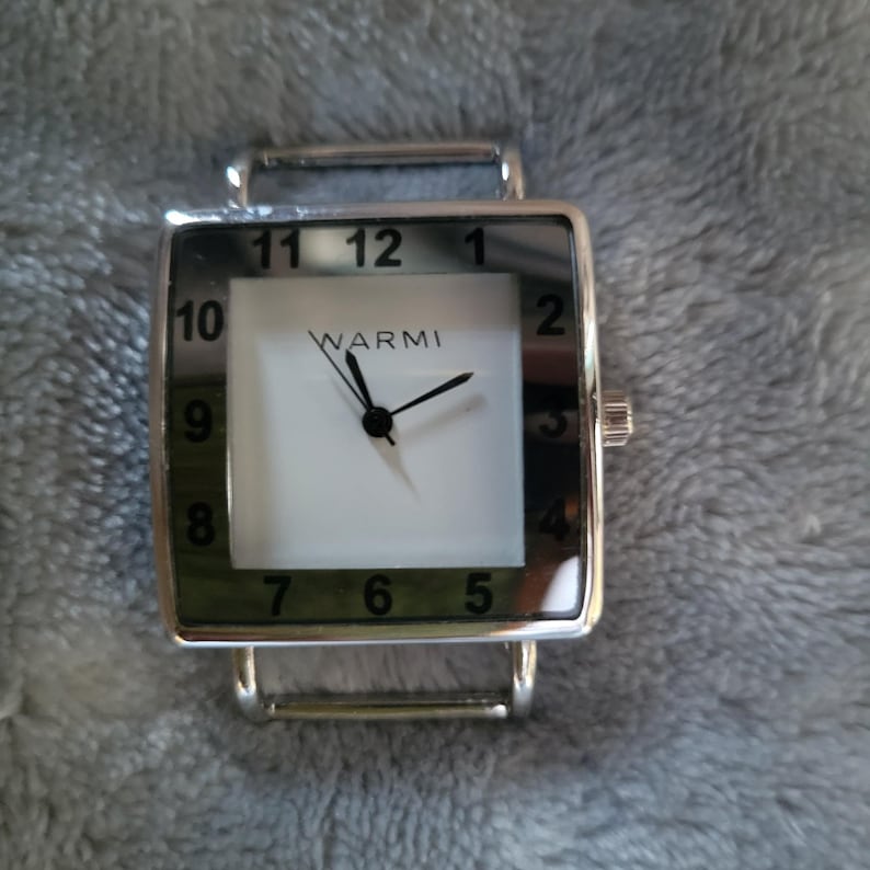 May include: A silver square watch with a white face and black numbers. The watch has a green border around the face and the word "WARMI" printed on the face.