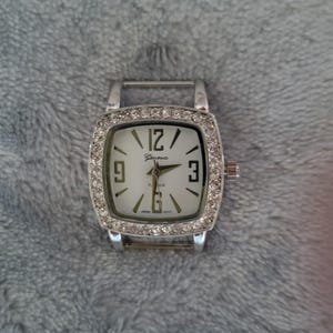 May include: A silver watch with a square face and a white dial. The watch is decorated with rhinestones around the bezel. The watch face has black Roman numeral hour markers and the word "Geneva" printed on it.