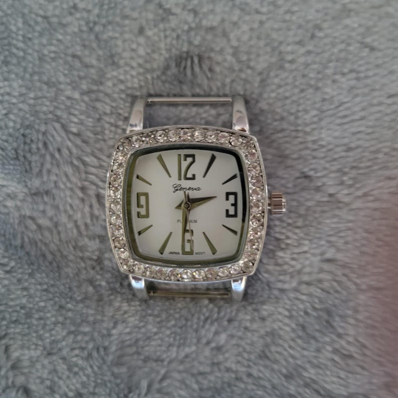 May include: A silver watch with a square face and a white dial. The watch is decorated with rhinestones around the bezel. The watch face has black Roman numeral hour markers and the word "Geneva" printed on it.