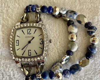 May include: A Geneva watch with a square, crystal-studded face and a white dial. The watch has silver-tone metal accents and is attached to a double-strand bracelet made of blue and silver beads. The watch is displayed on a white surface.