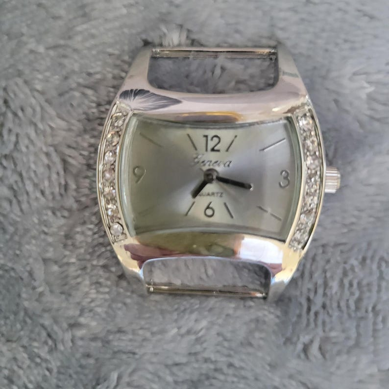 May include: A silver watch with a white face and a silver band. The watch has a crystal-encrusted bezel and the words "Geneva Quartz" on the face.