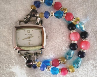 May include: A Geneva quartz watch with a silver rectangular face and a white dial. The watch is attached to a double-strand bracelet made of colorful beads in shades of blue, pink, yellow, and black. The watch face displays the time.