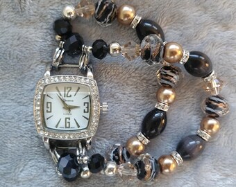 May include: A silver-toned Geneva Platinum watch with a square face, surrounded by rhinestones. The watch is paired with a beaded bracelet featuring black, gold, and clear beads. The watch face displays the numbers 12, 3, 6, and 9.