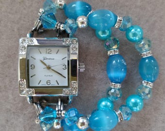 May include: A silver square watch with a white face and blue crystal accents. The watch is attached to a blue beaded bracelet.