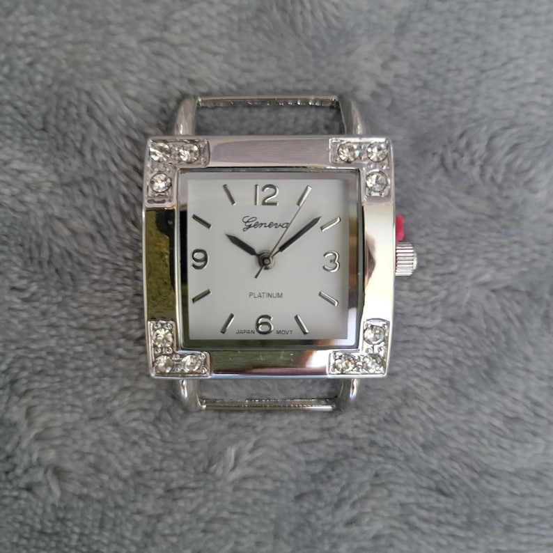 May include: A silver square watch with a white face and rhinestones around the bezel. The watch face has black hands and black Roman numeral hour markers. The watch face also has the words "Geneva" and "Platinum" printed on it.