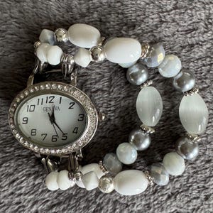 May include: A silver watch with a white face and crystal accents. The watch is attached to a bracelet made of white and gray beads.