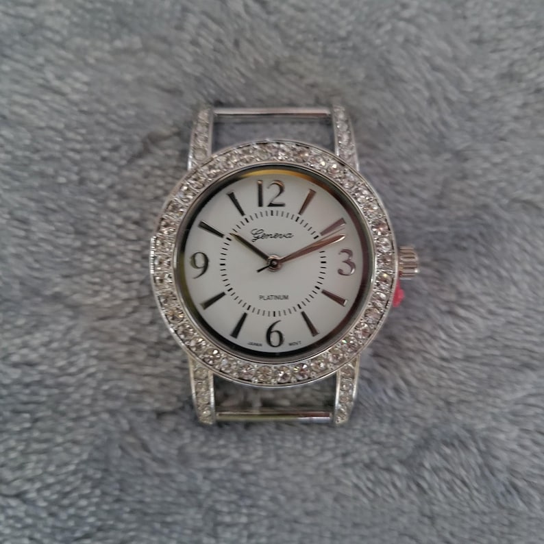 May include: A silver oval-shaped watch with a white face and a diamond-like bezel. The watch face has black Roman numeral hour markers and black hands. The watch face also has the word "Geneva" and "Platinum" printed on it.