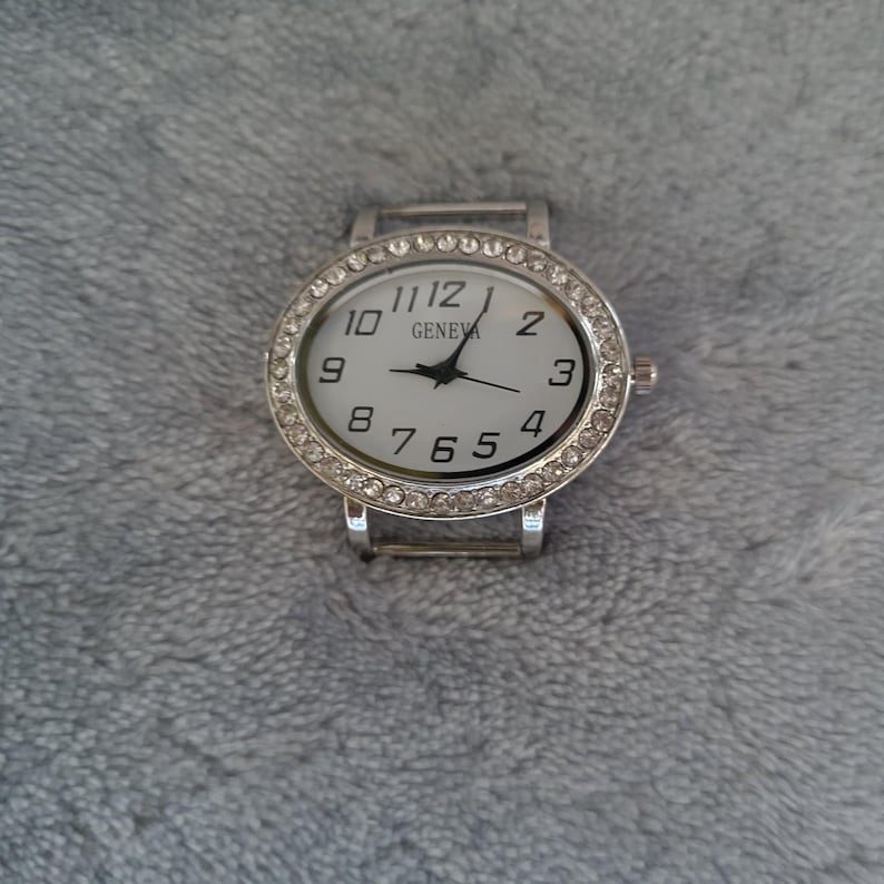 May include: A silver wristwatch with a white face and black numbers. The watch has a silver band and is decorated with rhinestones around the face. The face of the watch says "GENEVA" at the top.