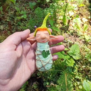 Puede incluir: Un pequeño gnomo de fieltro verde con un sombrero verde y un corazón de hoja en el pecho. El gnomo está envuelto en un paño verde y atado con cuerda. El gnomo está siendo sostenido en una mano contra un fondo verde de hojas.