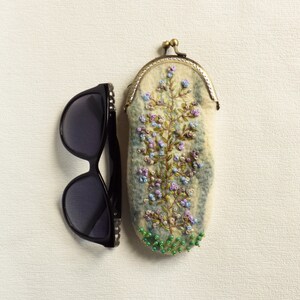 May include: A white felt eyeglass case with a floral embroidery design in shades of blue, green, and purple. The case has a brass clasp and is decorated with green beads.