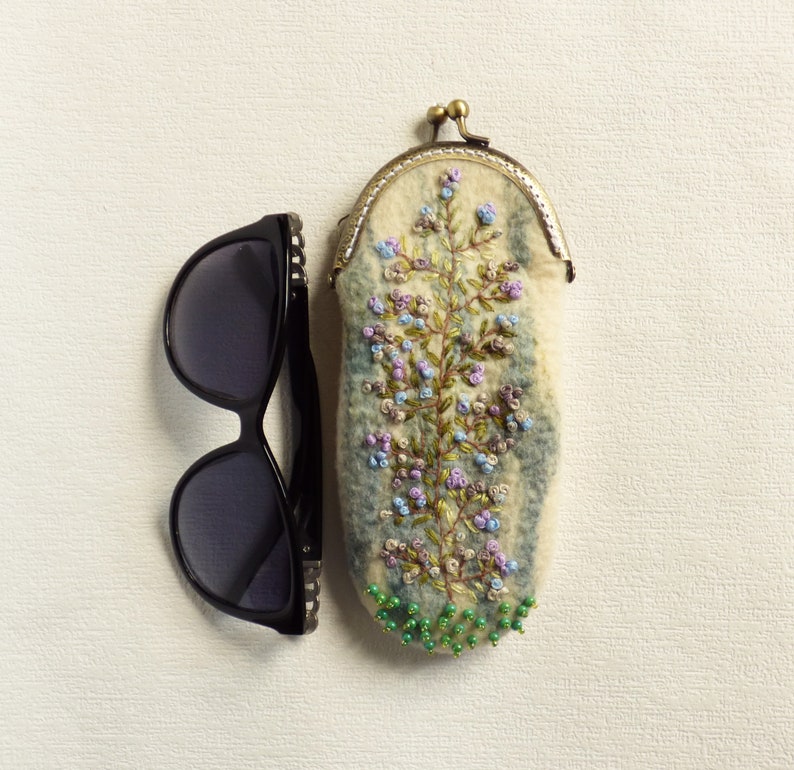 May include: A white felt eyeglass case with a floral embroidery design in shades of blue, green, and purple. The case has a brass clasp and is decorated with green beads.