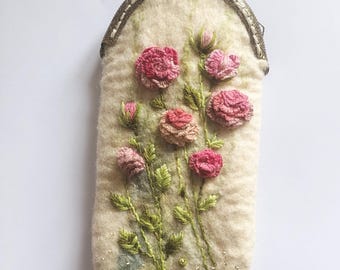 May include: A cream-coloured felted pouch with a bronze-toned metal kiss-lock clasp. The front features embroidered pink roses, green stems, and leaves, accented with small green beads at the base of the floral arrangement.
