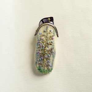 May include: A white felt eyeglass case with a floral embroidery design in shades of blue, green, and purple. The case has a brass clasp and is decorated with green beads.