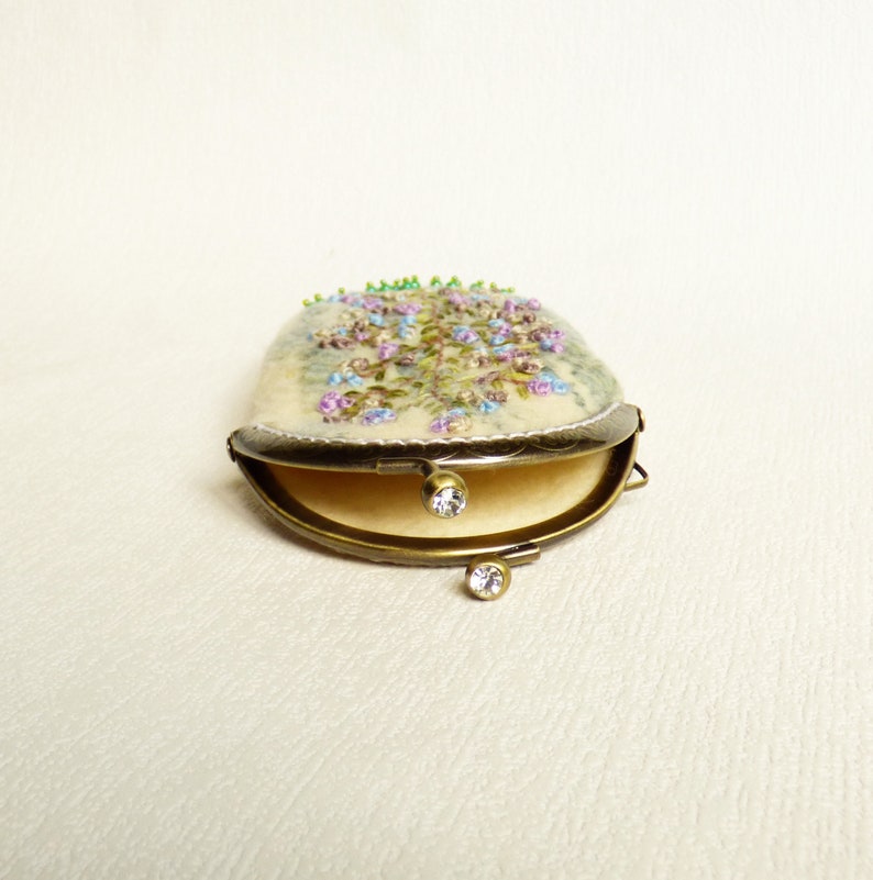 May include: A small, cream-coloured pouch with a floral embroidery design. The pouch is open and has a brass clasp with two clear rhinestones.